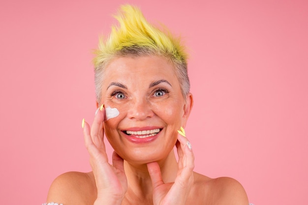 Femme mûre avec des cheveux teints en jaune élégants et des épaules nues appliquant une crème anti-âge sur fond de studio rose gymnastique faciale anti-âge