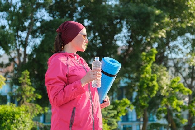Femme mûre en coupe-vent de sport avec une bouteille d'eau de tapis de yoga, marchant en plein air. Mode de vie sain et actif, sport, forme physique chez les personnes d'âge moyen