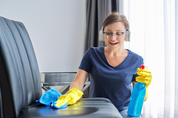 Femme mûre dans des gants de casque avec un détergent de chiffon nettoyant à la maison