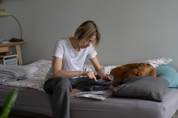 Femme mûre à l'expression épuisée assise sur le lit avec un chien vizsla compte les factures d'électricité