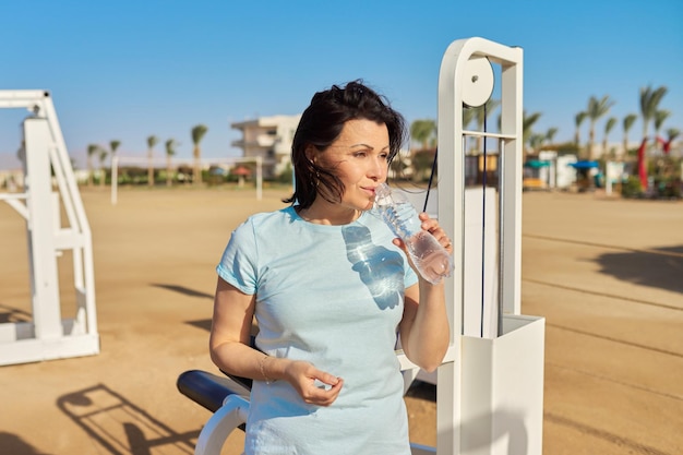 Femme mûre faisant des exercices sportifs sur des simulateurs extérieurs avec une bouteille d'eau