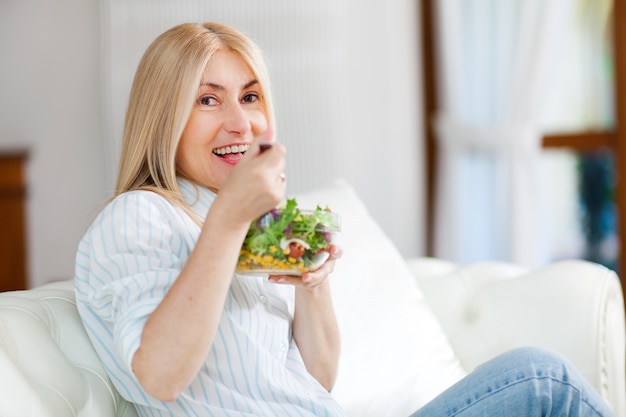 Femme mûre, manger une salade sur son canapé