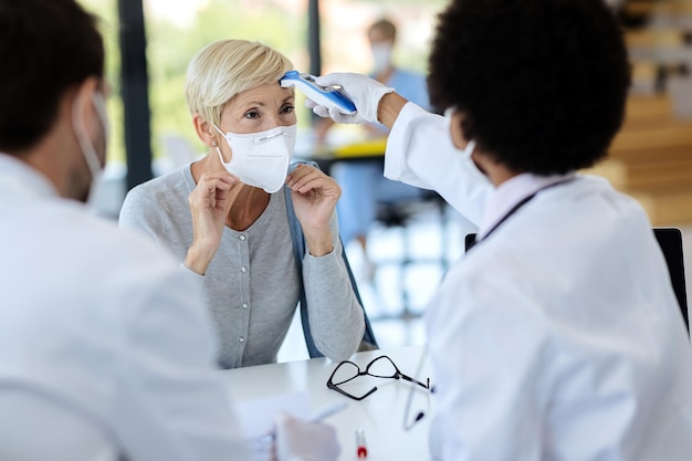 Femme mûre avec masque facial faisant mesurer sa température lors d'un rendez-vous médical