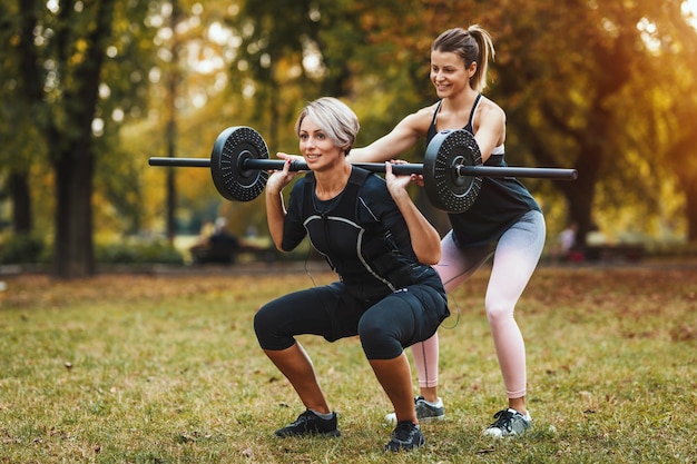 Une femme mûre musclée fait des exercices de squat avec un entraîneur personnel dans le parc, vêtue d'un costume noir avec un simulateur électronique EMS pour stimuler ses muscles.