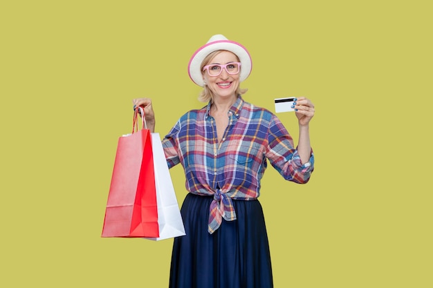Femme mûre positive souriante en chapeau et lunettes tenant un sac à provisions et montrant une carte de crédit