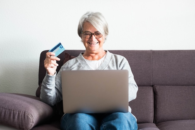 Une femme mûre s'amusant seule à la maison avec sa carte de crédit et son ordinateur portable faisant des achats en ligne sur le canapé - accro du shopping senior souriant et regardant l'ordinateur