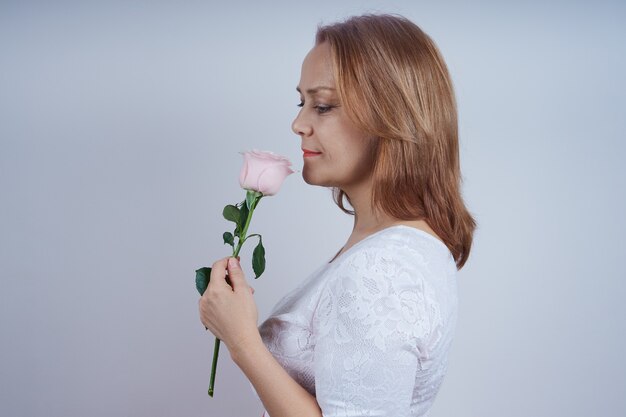 Une femme mûre se tient de profil, dans une robe blanche tenant une fleur rose. dans le mur gris.