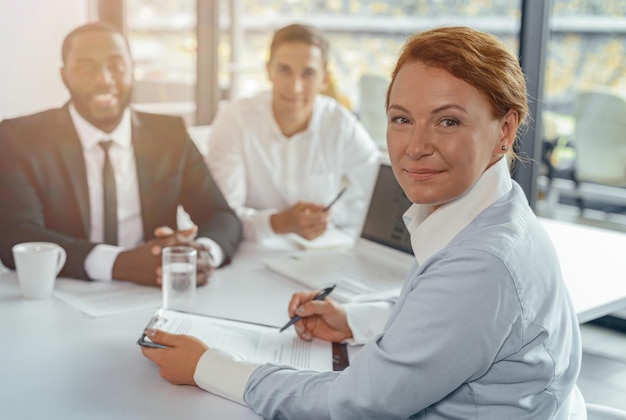 Femme mûre souriante en vêtements d'affaires regardant la caméra assise à table au bureau avec une tablette et un stylo dans les mains se préparant à signer un contrat