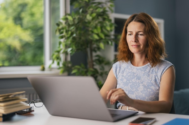 Femme mûre en tenue décontractée assise au bureau et travaillant sur un ordinateur portable sans fil Belle dame utilisant un ordinateur portable dans un salon lumineux