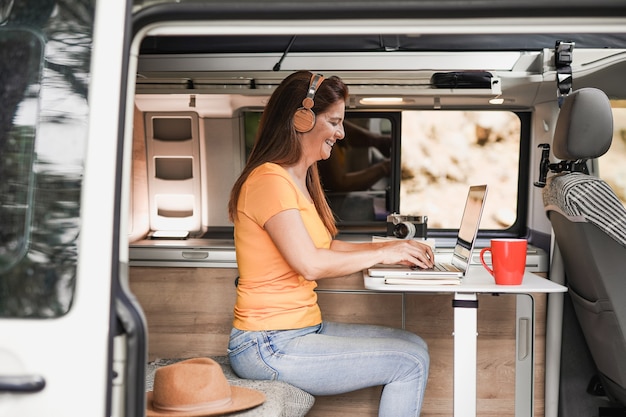 Femme mûre travaillant à l'intérieur d'un mini van camper avec ordinateur portable portant un casque sans fil