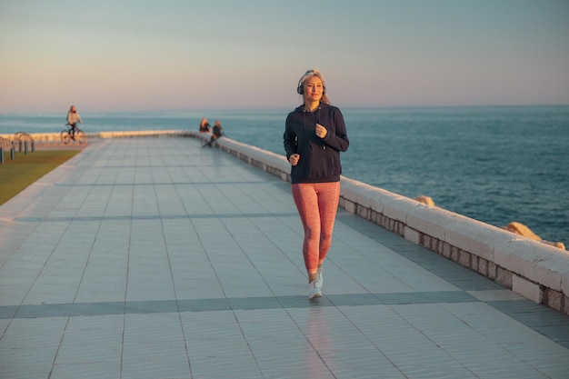 Femme mûre en vêtements de sport courant le long du bord de la mer le soir