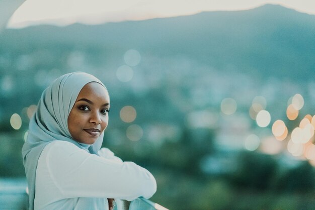 Femme musulmane africaine dans la nuit sur un balcon souriant à la caméra avec des lumières bokeh de la ville en arrière-plan. Photo de haute qualité