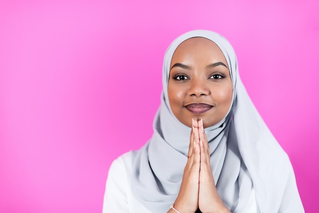 La femme musulmane africaine moderne fait la prière traditionnelle à Dieu, garde les mains dans le geste de prière, porte des vêtements blancs traditionnels, a une expression faciale sérieuse, isolée sur fond rose en plastique