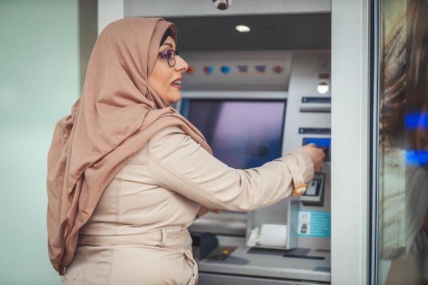 Une femme musulmane d'âge moyen portant le hijab se tient près d'un distributeur automatique de billets dans un environnement urbain, tirant de l'argent ou vérifiant le solde du compte.
