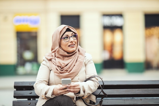 Une femme musulmane d'âge moyen portant le hijab avec un visage heureux est assise sur le banc en milieu urbain, renvoyant des messages sur son smartphone.