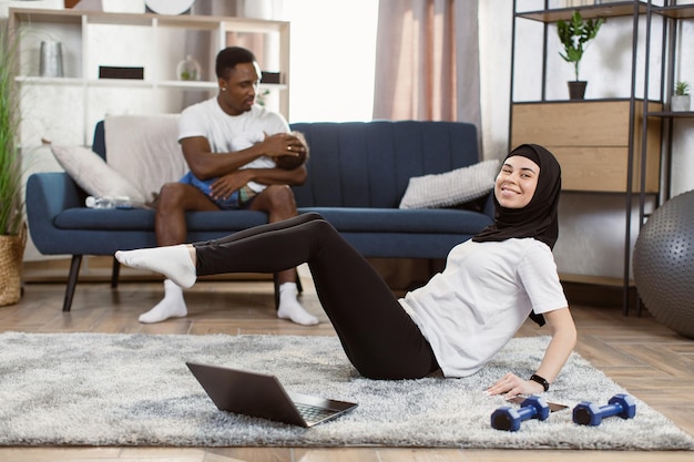 Une femme musulmane arabe dans un legging de sport et un t-shirt blanc pratique le yoga dans le salon