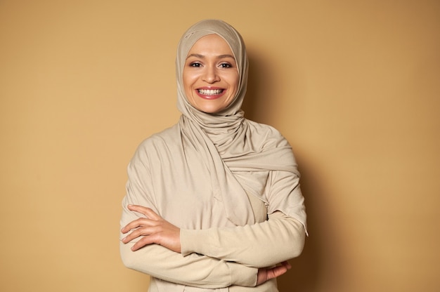 Femme musulmane arabe souriante posant avec les bras croisés sur une surface beige avec espace de copie