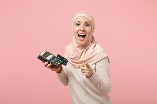 Femme musulmane arabe en vêtements légers hijab posant isolé sur fond rose. Concept de style de vie religieux des gens. Maquette de l'espace de copie. Tenir le terminal de paiement pour traiter, acquérir les paiements par carte de crédit.