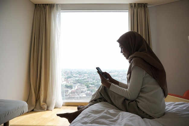 Femme musulmane asiatique assise sur le lit de la chambre le matin pour profiter de la vue extérieure tout en utilisant un téléphone portable