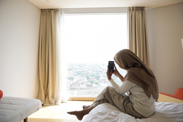 Femme musulmane asiatique assise sur le lit de la chambre le matin pour profiter de la vue extérieure tout en utilisant un téléphone portable