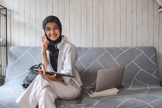 Femme musulmane asiatique assise et tenant des livres tout en ayant un appel téléphonique à la maison