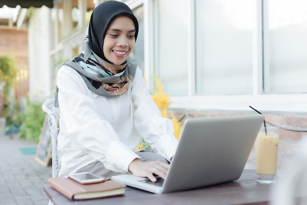 Une femme musulmane asiatique est assise dans un jardin avec un ordinateur portable et un téléphone sur ses genoux