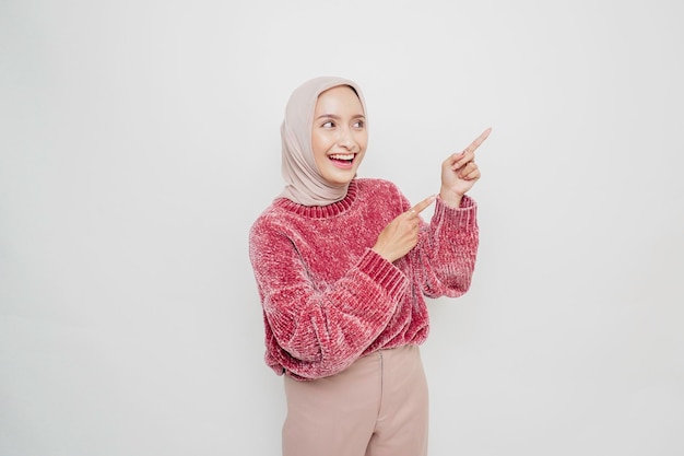Femme musulmane asiatique excitée portant un pull rose et un hijab pointant vers l'espace de copie à côté d'elle isolé sur fond blanc
