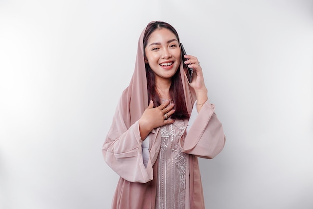 Une femme musulmane asiatique heureuse portant un foulard parlant joyeusement au téléphone isolée par fond blanc