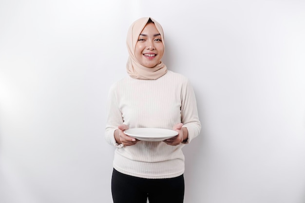 Une femme musulmane asiatique jeûne et a faim et tient une assiette tout en regardant de côté en pensant à quoi manger