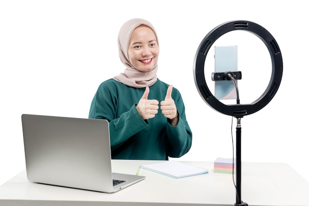 Une femme musulmane asiatique avec un ordinateur portable et des téléphones portables avec un équipement d'éclairage fait un vlog isolé sur fond blanc