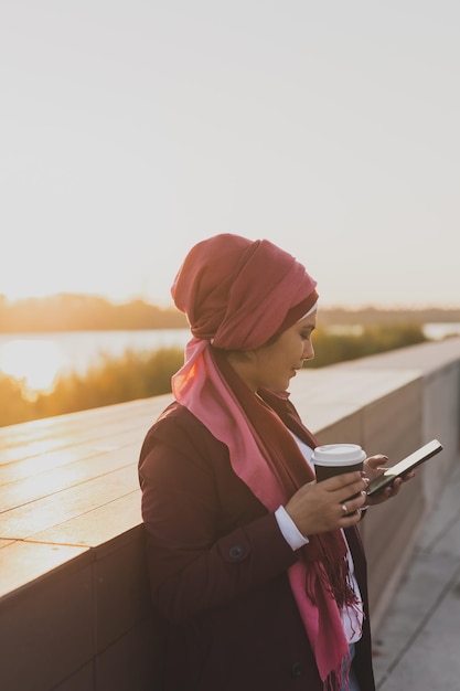 Femme musulmane asiatique portant le hijab faisant défiler les réseaux sociaux ayant une pause-café en plein air sur fond de ville