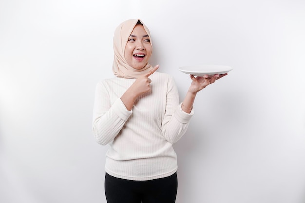 Une femme musulmane asiatique souriante jeûne et a faim et tient et pointe vers une assiette