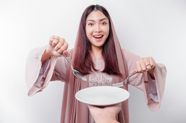 Une femme musulmane asiatique souriante jeûne et a faim et tient et pointe vers une assiette