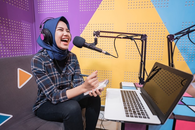 Une femme musulmane excitée enregistre un podcast en studio avec un ordinateur portable