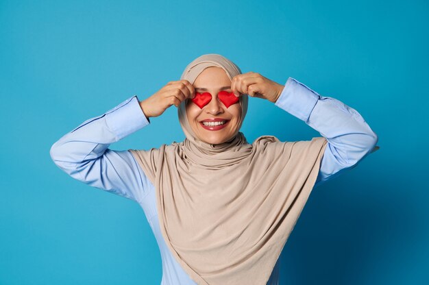 Femme musulmane en hijab avec apparence orientale mettant des formes de coeur sur ses yeux et souriant avec un sourire à pleines dents.