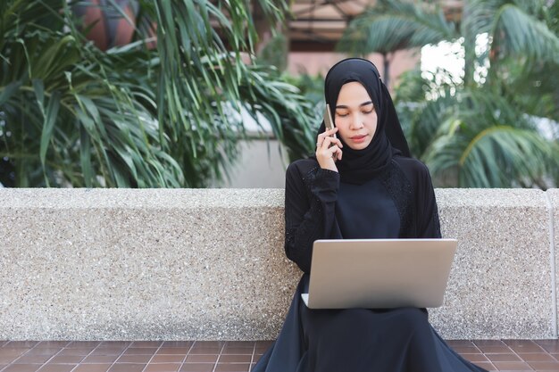 Femme musulmane en hijab noir parler téléphone et travailler avec un bureau extérieur pour ordinateur portable.