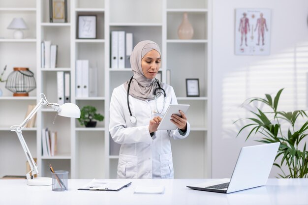 Une femme musulmane en manteau médical blanc se tient à l'intérieur du bureau médical d'une clinique.