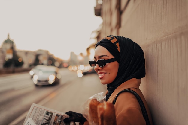 Une femme musulmane moderne avec un hijab se promène dans la ville tout en portant un journal et du pain à la main.