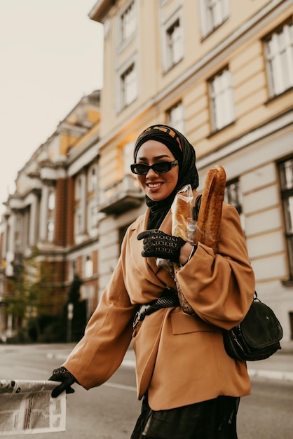 Photo une femme musulmane moderne avec un hijab se promène dans la ville tout en portant un journal et du pain à la main.