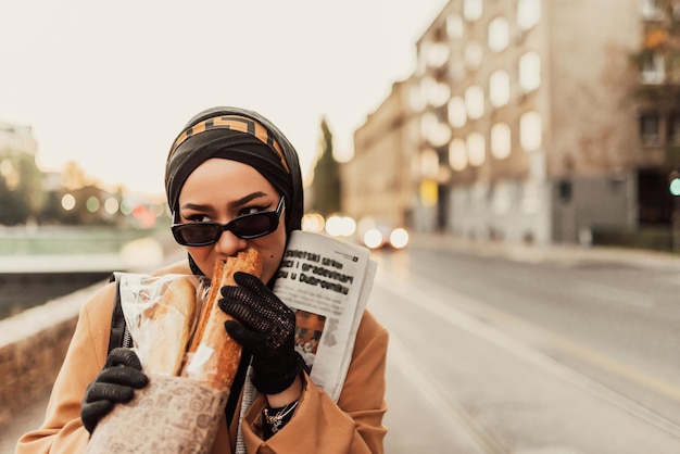 Une femme musulmane moderne avec un hijab se promène dans la ville tout en portant un journal et du pain à la main.