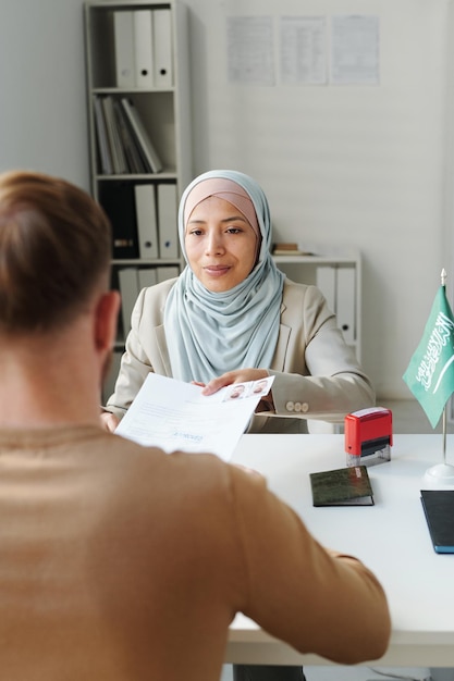 Femme musulmane moderne travaillant à l'ambassade assise au bureau devant un homme lui passant des documents après un