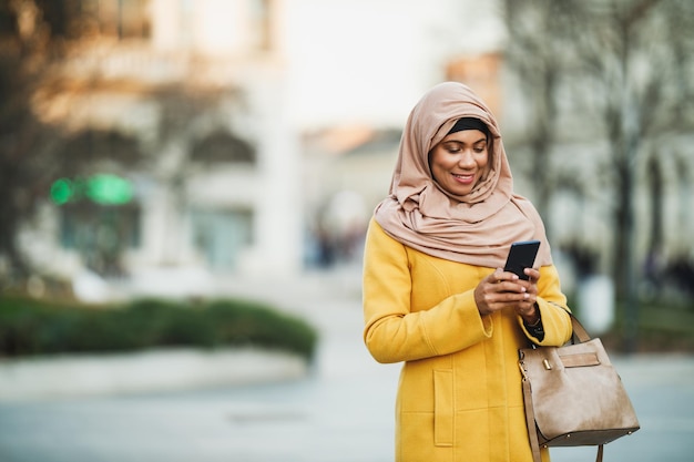 Femme musulmane noire souriante portant un hijab et renvoyant des messages sur son smartphone tout en marchant dans un environnement urbain.