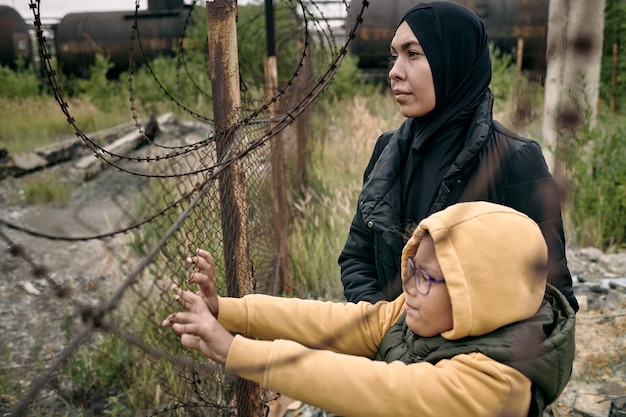 Femme musulmane pleine d'espoir en hijab debout avec sa fille au fil de fer barbelé et regardant à distance