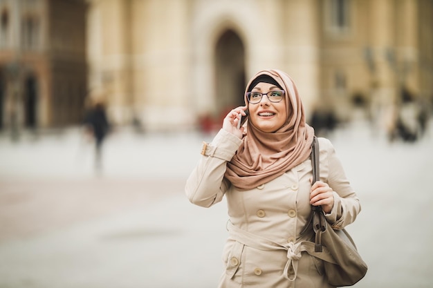 Femme musulmane souriante portant un hijab et parlant sur son smartphone tout en marchant en milieu urbain.
