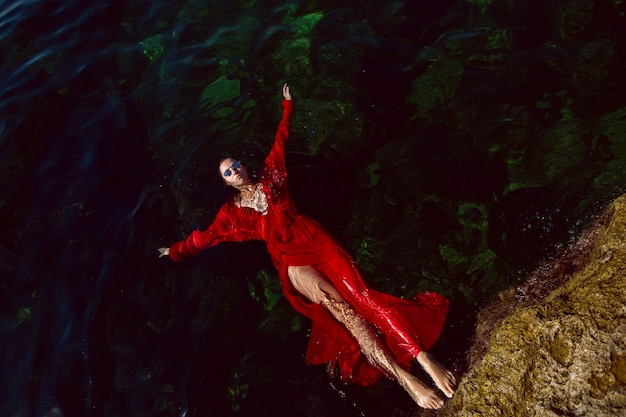 Femme nage dans la mer dans une robe longue rouge avec des lunettes de soleil