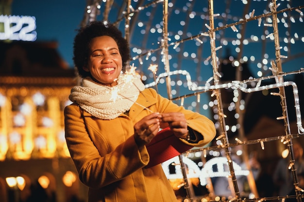 Femme noire d'âge moyen tenant des cierges magiques et un cadeau de Noël lors d'une nuit d'hiver festive dans la ville.