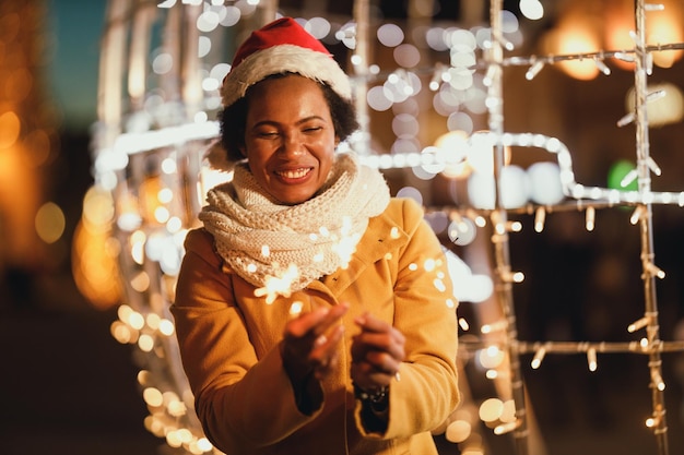 Femme noire d'âge moyen tenant des cierges magiques lors d'une nuit d'hiver festive dans la ville.