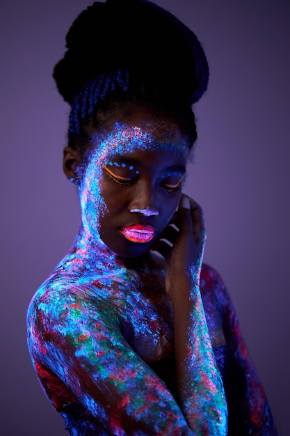 Femme noire avec art corporel brillant dans la lumière ultraviolette. belle femme peinte en poudre fluorescente, se touche. espace violet foncé isolé. beauté, concept de mode. tournage inhabituel