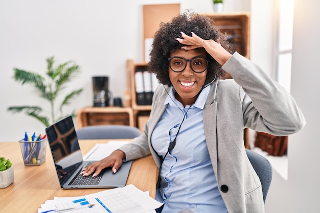 Femme noire aux cheveux bouclés portant un casque d'agent de centre d'appel au bureau très heureuse et souriante regardant loin avec la main sur la tête. notion de recherche.