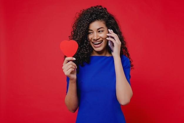 Femme noire avec carte de Saint Valentin en forme de coeur parler au téléphone sur le mur rouge